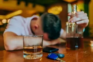 young man rests head on bar. An unfinished drink, keys and bottle rest on the bar in the foreground.